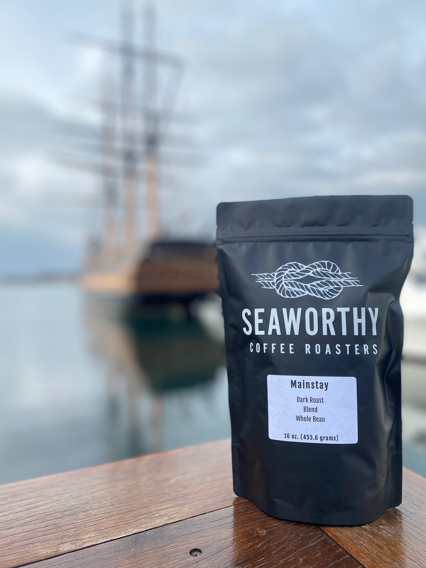 Seaworthy slow roasted, small batch, low acid coffee. 1 pound bag of Mainstay dark roast specialty coffee. Bag of coffee on Bowens Wharf in Newport Rhode Island. Old ship with three masts in background.