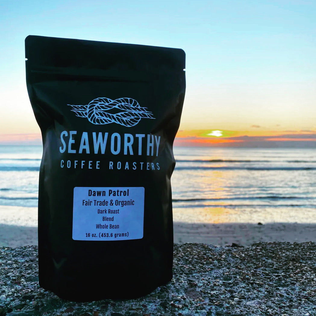 Seaworthy slow roasted, small batch, low acid coffee. 1 pound bag of Dawn Patrol dark roast specialty coffee blend.  Bag of coffee on beach.  Sunrise over the ocean.